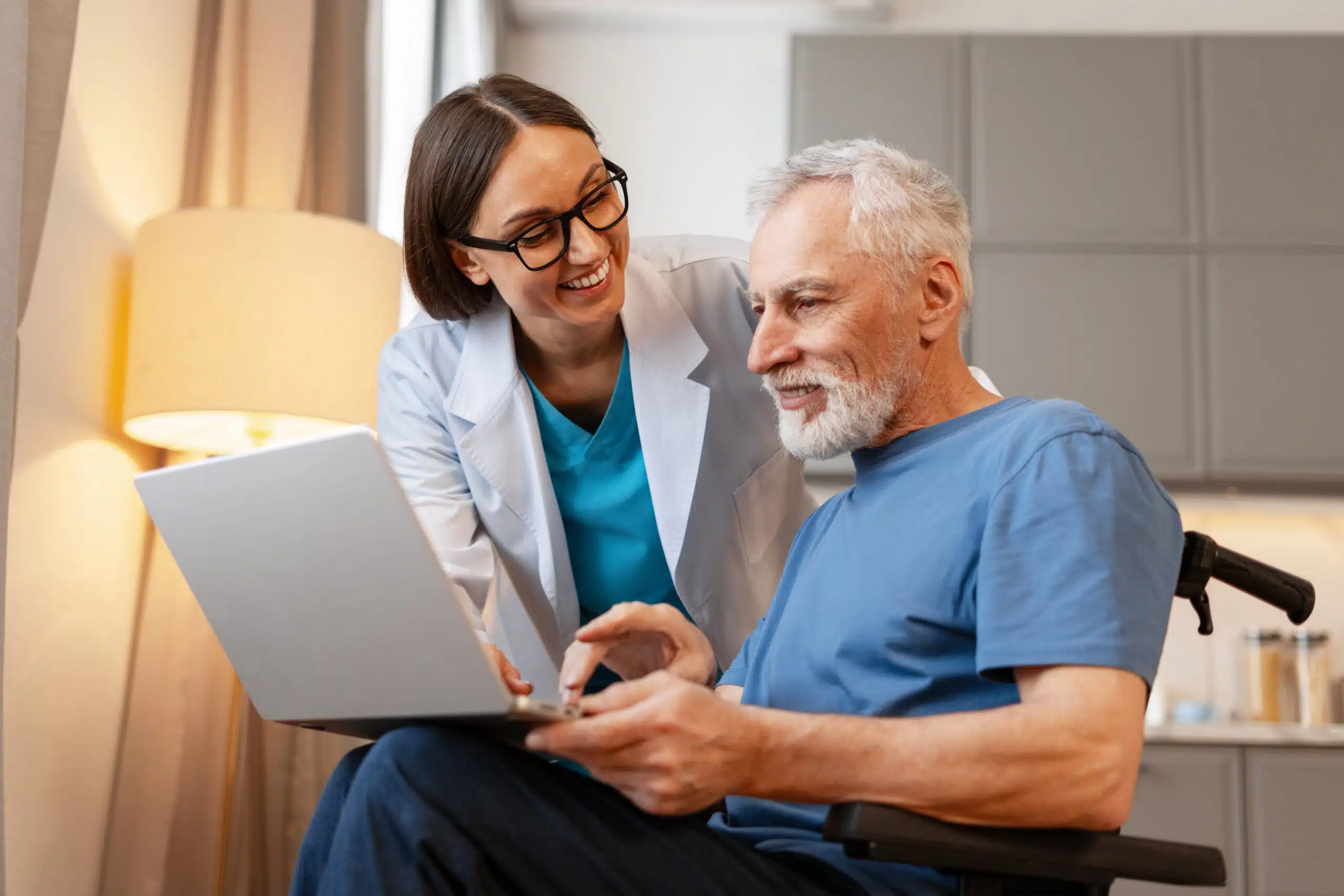 What To Do Before and After an IME Appointment 1 Smiling doctor helping a happy senior man in a wheelchair using a laptop, providing medical and technological support in a home healthcare setting
