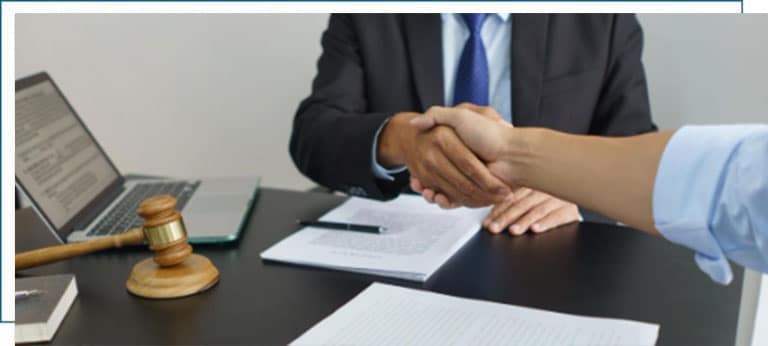 Two people shaking hands across a desk with legal documents, a laptop, and a judge’s gavel, symbolizing a legal agreement or settlement.