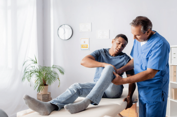 A physical therapist in scrubs examining a patient’s injured knee as the patient winces in pain while sitting on a treatment table in a medical office.
