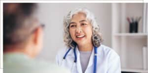 Smiling female doctor with gray curly hair wearing a white coat and stethoscope, speaking with a patient in a medical office.