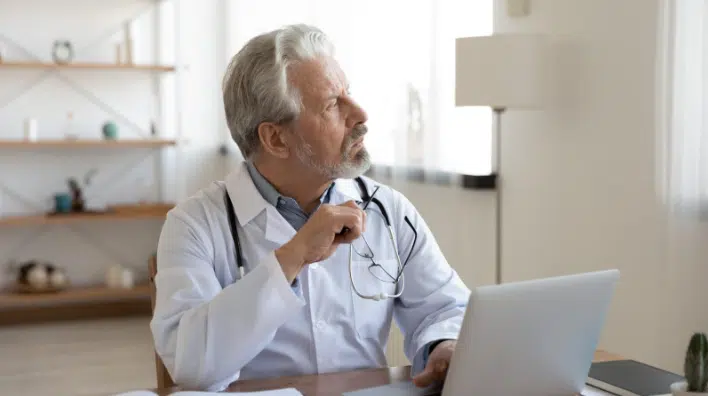 Older male doctor with gray hair and beard, holding eyeglasses and looking thoughtfully out a window while seated at a desk with a laptop.