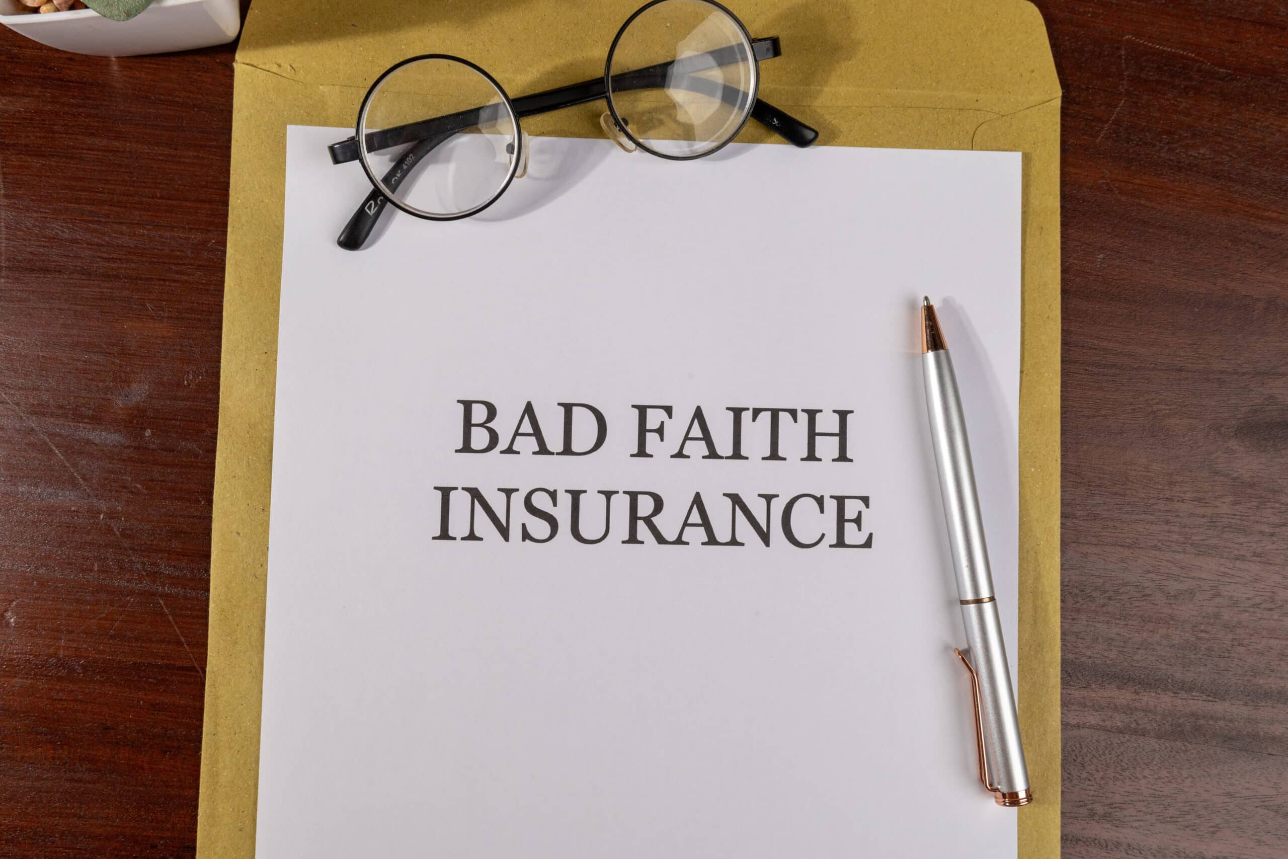Document labeled "Bad Faith Insurance" on a manila envelope with round-frame glasses and a silver pen on a wooden desk