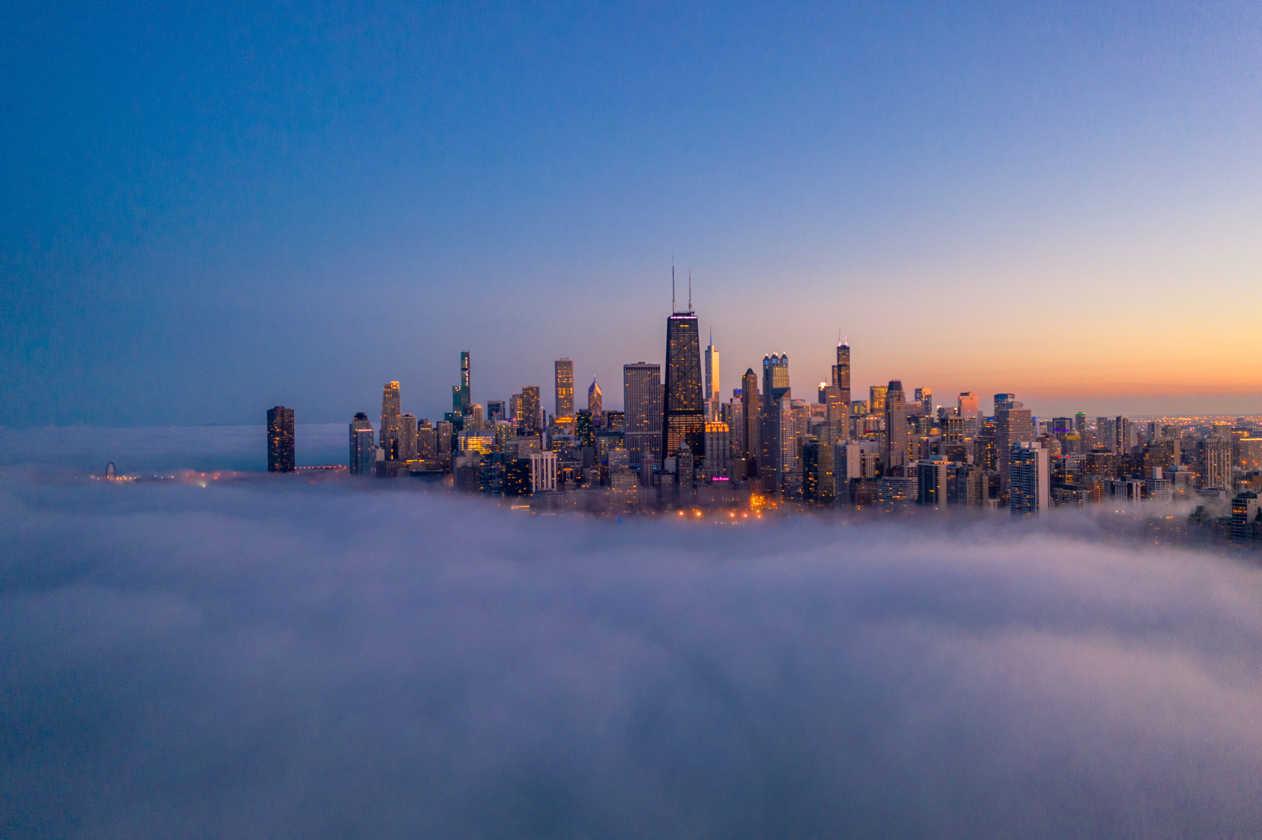 Chicago skyline emerging above a thick layer of fog at dusk, with illuminated skyscrapers against a blue and orange twilight sky