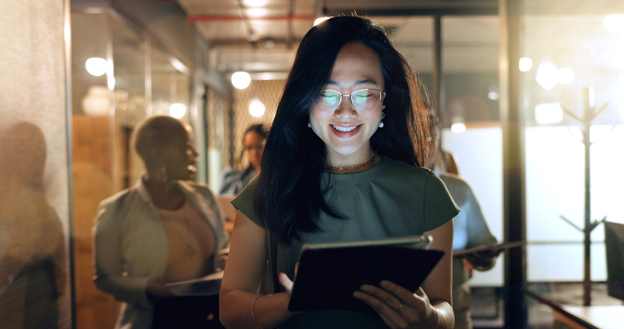 Professional woman wearing glasses reviews a tablet in a dimly lit office hallway with colleagues in the background