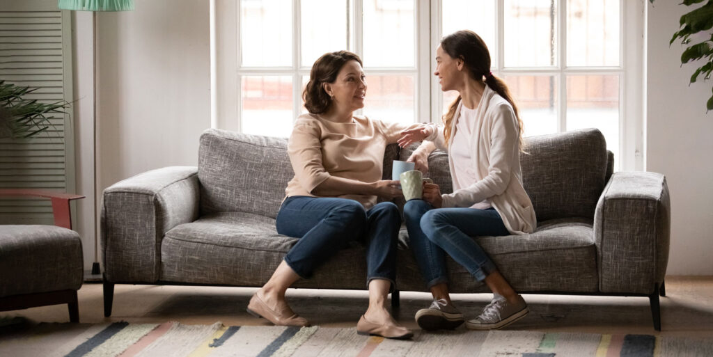 Two women sitting on a gray couch having a supportive conversation over coffee in a bright, comfortable living room