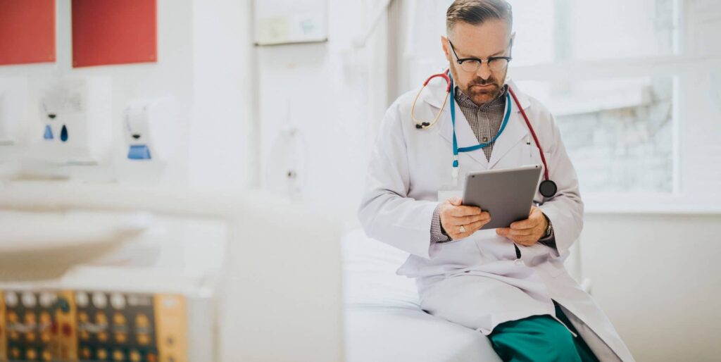 Male physician in white coat and stethoscope reviewing patient records on a tablet while seated in a medical exam room