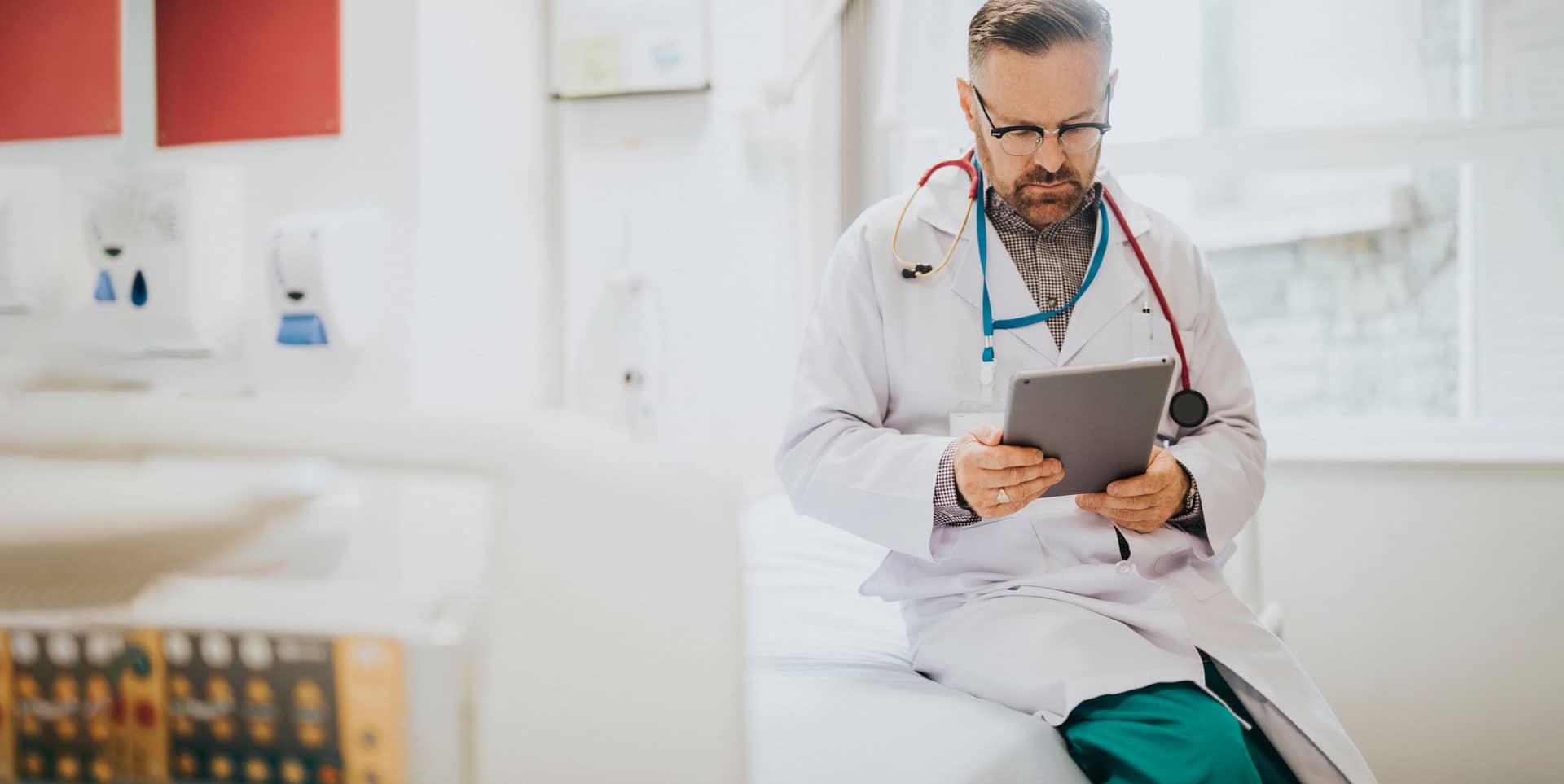 Male physician in white coat and stethoscope reviewing patient records on a tablet while seated in a medical exam room