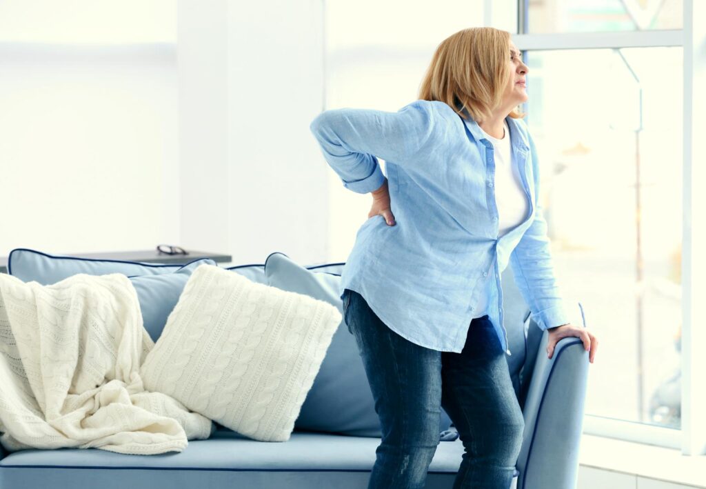 Middle-aged woman grimacing in lower back pain while rising from a couch, holding her back with one hand
