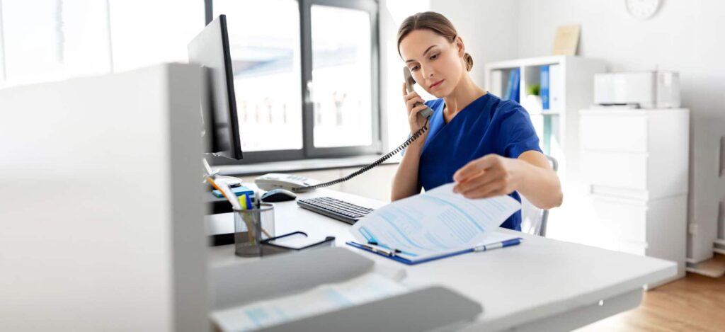 Healthcare professional in blue scrubs reviewing medical documents while on a phone call at an office desk with a computer
