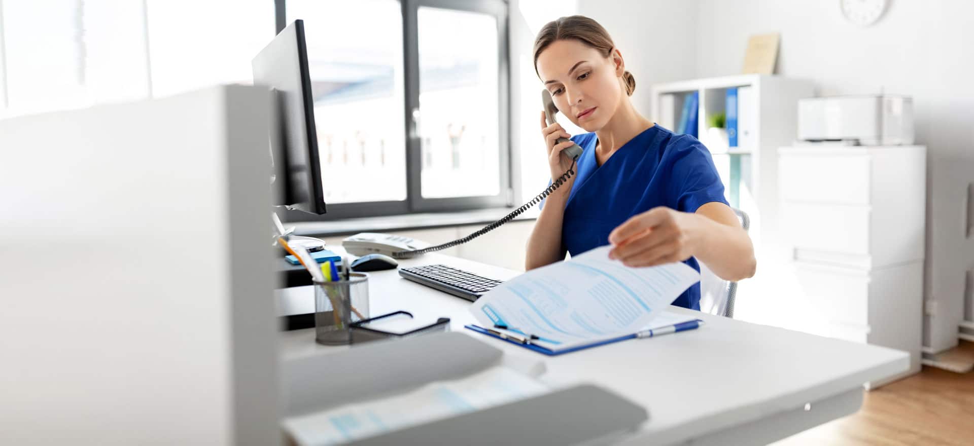 Healthcare professional in blue scrubs reviewing medical documents while on a phone call at an office desk with a computer