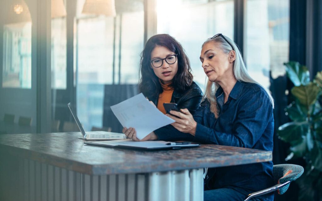 Two women reviewing legal documents and a smartphone together at a conference table in a modern office setting