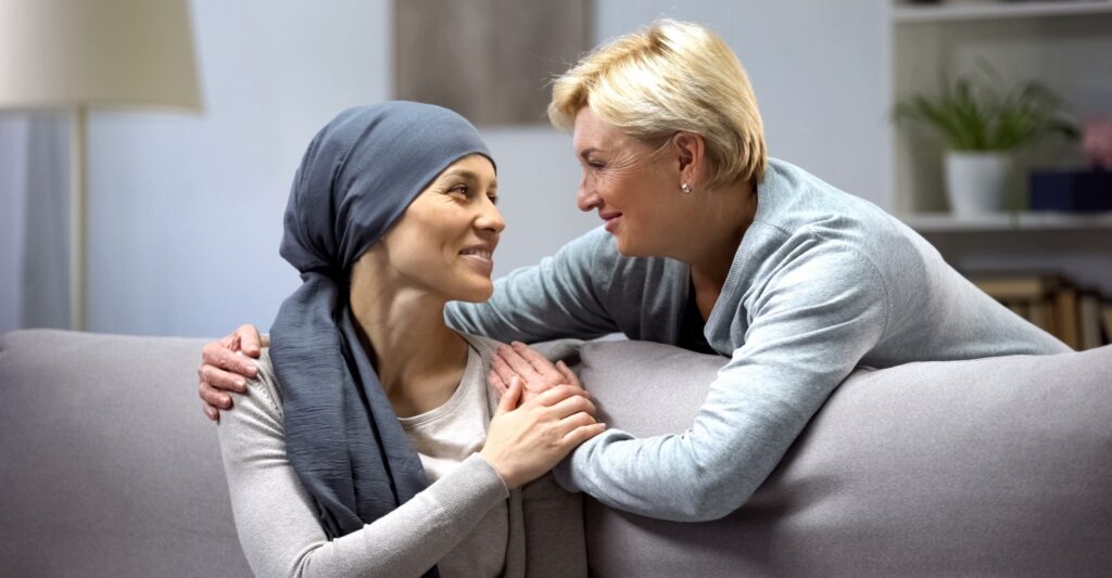 Supportive woman comforting a cancer patient wearing a head scarf, sharing an encouraging moment together on a couch