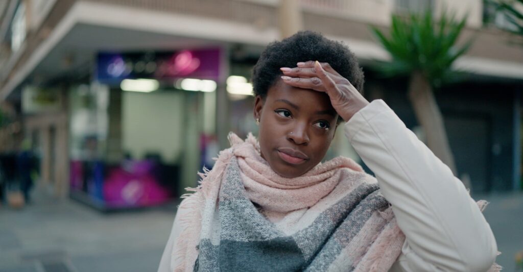 Woman holding her hand to her forehead in distress on a city street, wearing a scarf and white jacket