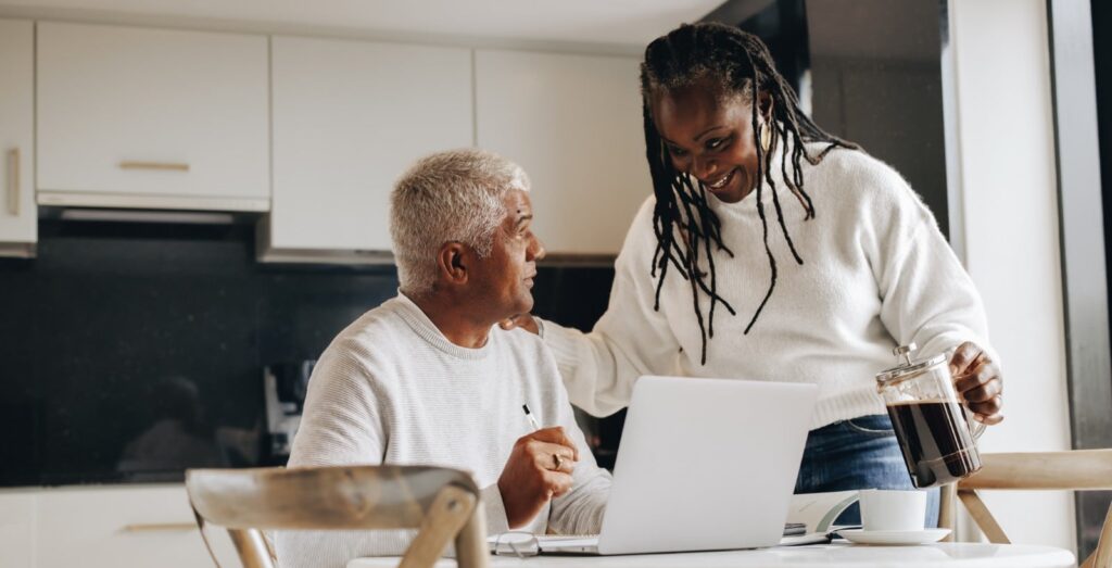 Middle-aged couple reviewing information on a laptop together at a kitchen table, sharing a warm, supportive moment over coffee