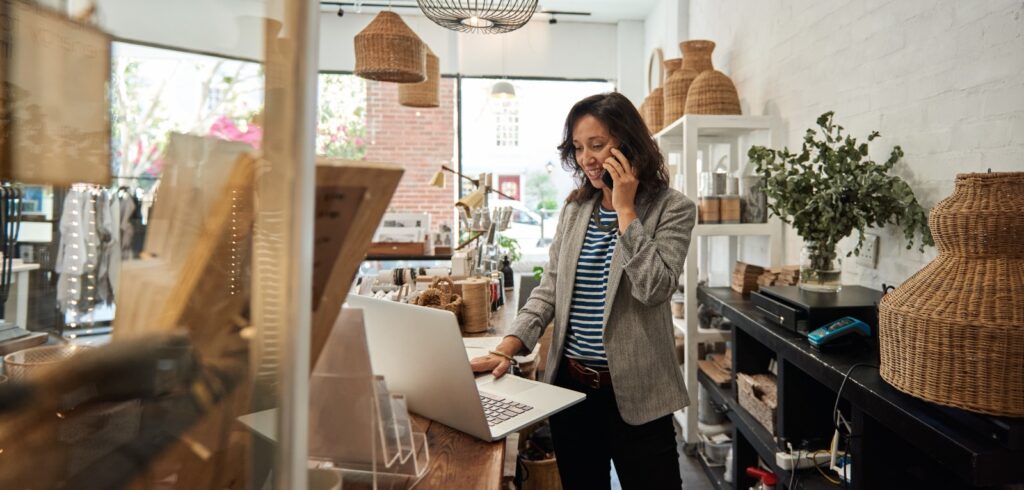 Small business owner talking on phone while working on laptop at boutique shop counter
