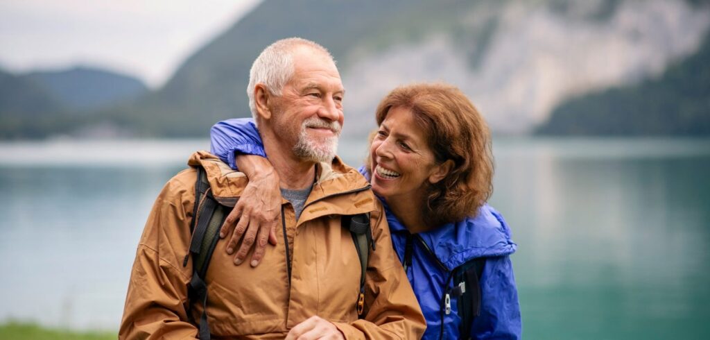 Smiling senior couple in hiking jackets embracing outdoors near a mountain lake, enjoying an active retirement lifestyle
