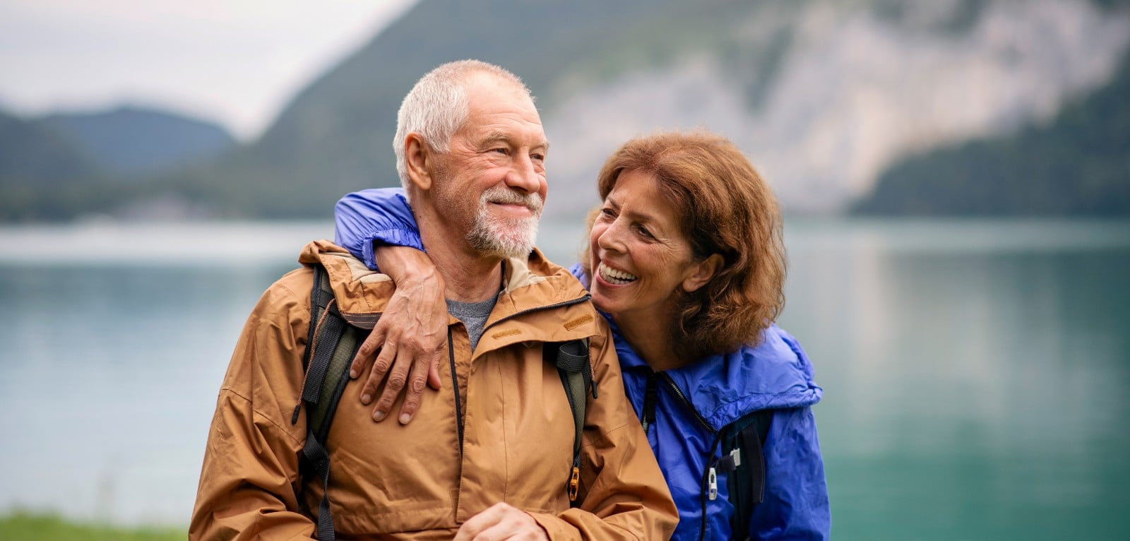 Smiling senior couple in hiking jackets embracing outdoors near a mountain lake, enjoying an active retirement lifestyle