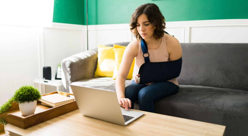 Woman with arm sling sitting on couch researching personal injury legal options on a laptop at home