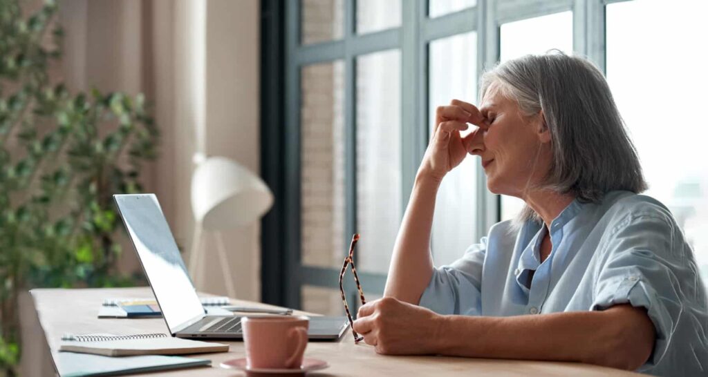 Stressed older woman holding glasses and pinching her nose at a desk with a laptop, notebook, and coffee cup nearby