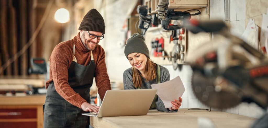 Two woodworking shop workers reviewing documents and a laptop together at a workbench surrounded by tools