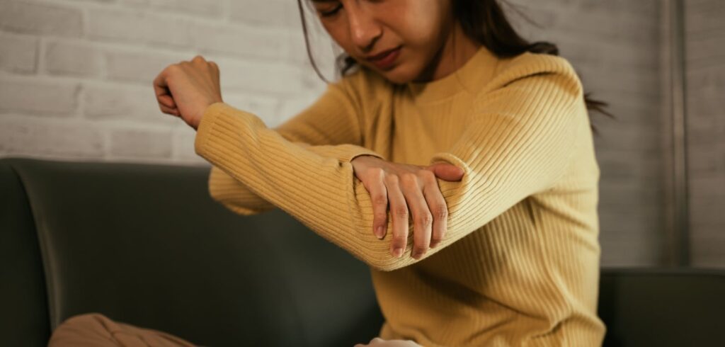 Woman in yellow sweater grimacing while holding her injured elbow in pain while seated on a couch