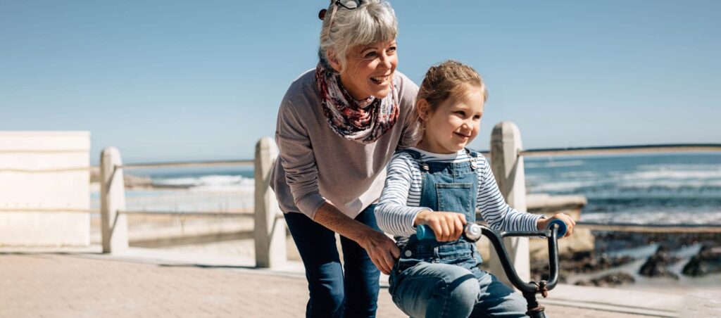 Grandmother helping young granddaughter ride a bicycle on a sunny seaside boardwalk, both smiling joyfully