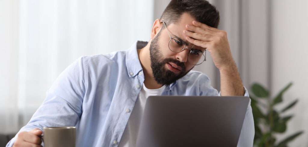 Stressed man holding coffee cup reads laptop screen with hand on forehead, showing concern or frustration