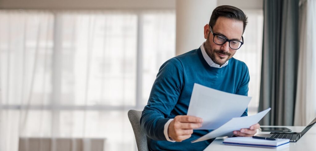 Man wearing glasses reviewing legal documents at a desk with a laptop in a bright professional setting