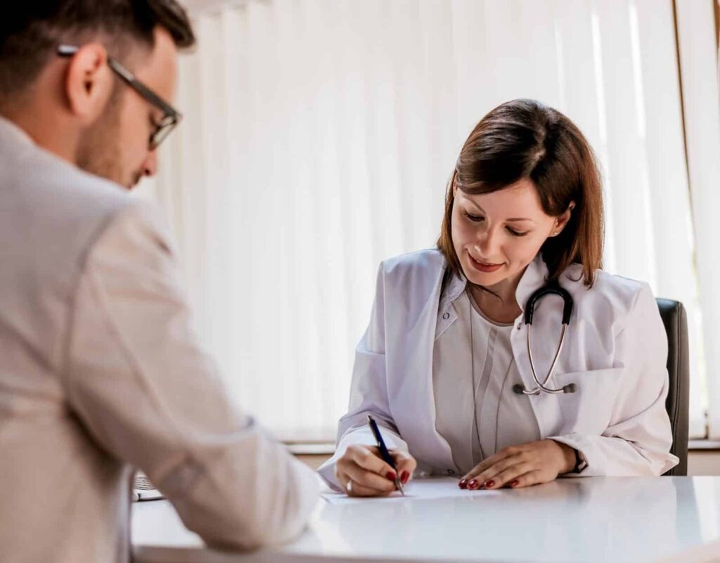Female doctor in white lab coat with stethoscope writing notes while consulting with a male patient at her desk