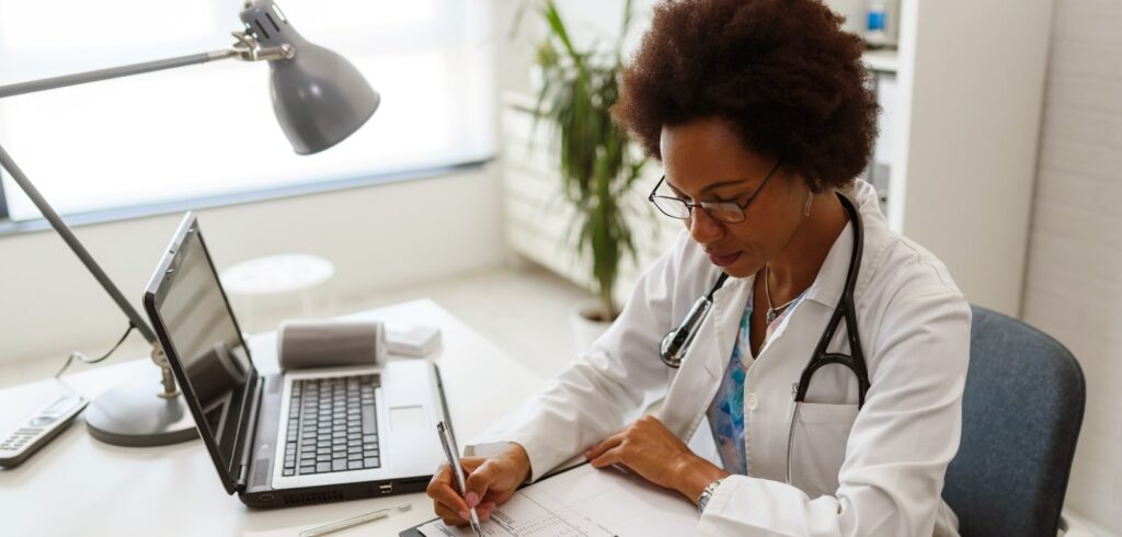 Female doctor in white coat and stethoscope writing medical notes at office desk with laptop