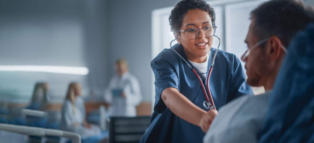 Nurse in blue scrubs with stethoscope attentively examining a patient in a hospital ward setting