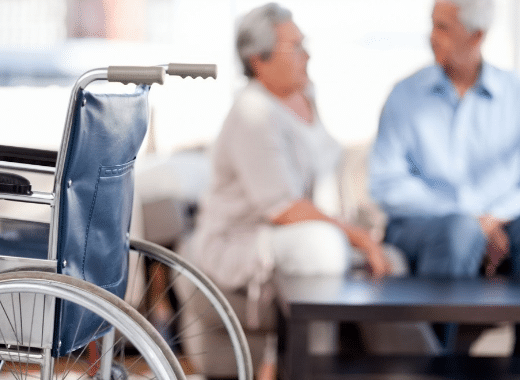 Wheelchair in foreground with two elderly adults seated and conversing in the blurred background