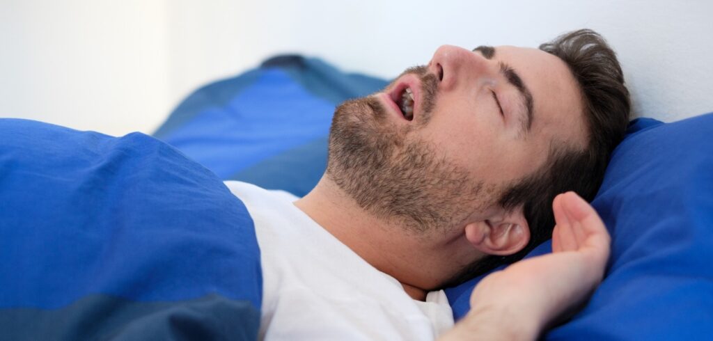 Man sleeping with mouth open on blue pillow, showing signs of snoring or sleep apnea