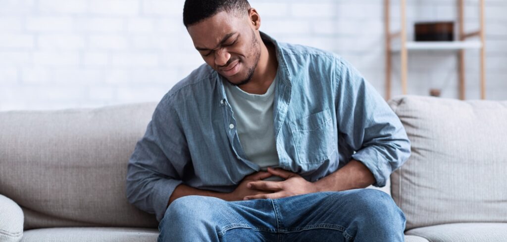 Man sitting on a couch clutching his stomach in pain, grimacing due to severe abdominal discomfort