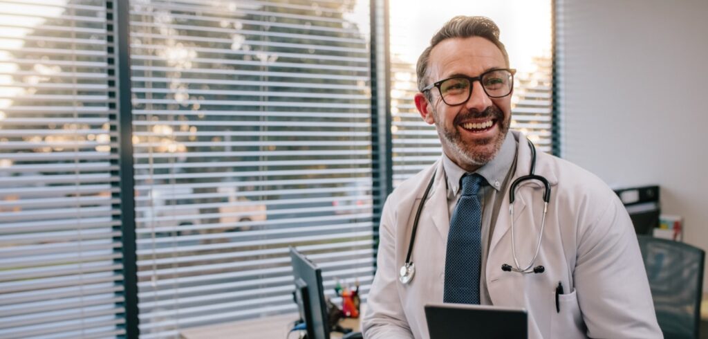 Smiling male doctor in white coat and stethoscope holding a tablet in a medical office with window blinds in background