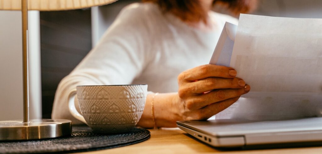 Woman reviewing legal documents at a desk with a laptop and coffee cup, examining paperwork from an envelope