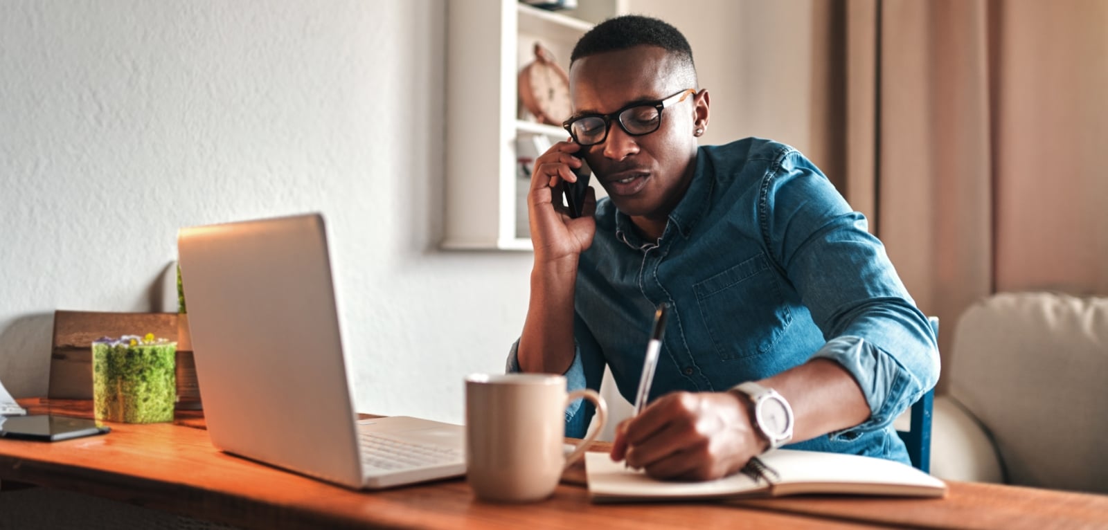 Man talking on phone while taking notes at a home desk with a laptop and coffee mug, consulting with a legal professional