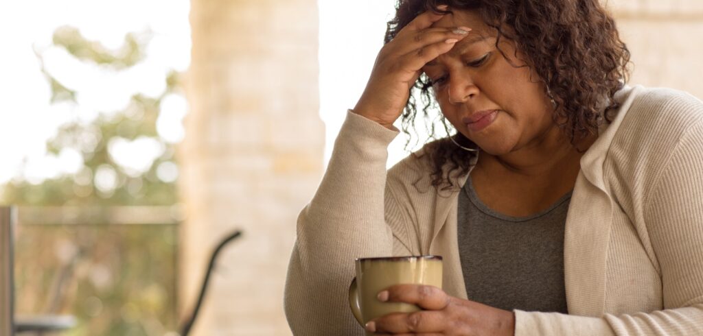 Woman holding a coffee mug with hand pressed to her forehead, appearing stressed or in pain outdoors