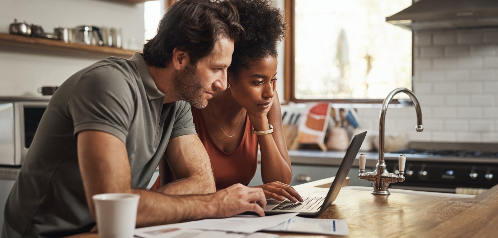 Couple reviewing legal documents together on a laptop at a kitchen counter with paperwork spread out before them