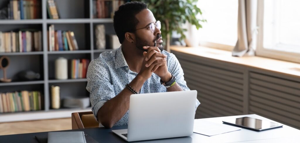 Man wearing glasses sits at desk with laptop, gazing thoughtfully toward window in a book-lined home office setting