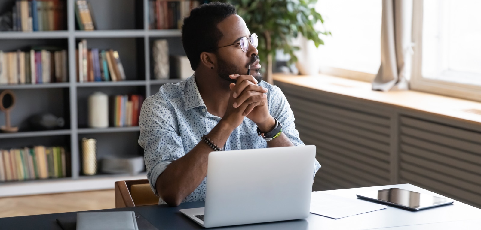 Man wearing glasses sits at desk with laptop, gazing thoughtfully toward window in a book-lined home office setting