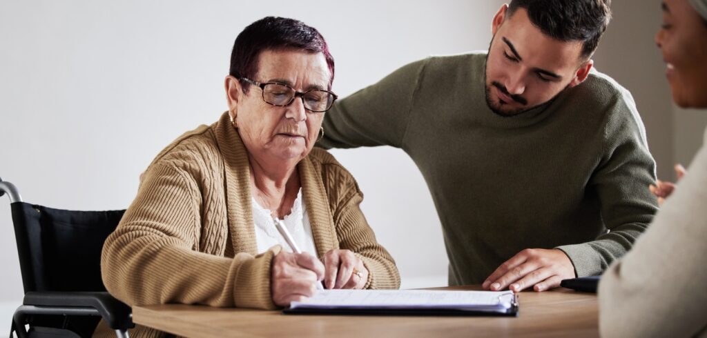 Elderly woman in wheelchair signing legal documents at a table while a young man assists her during a consultation