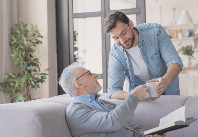 Young man handing a coffee mug to an elderly man resting on a couch at home, showing compassionate care