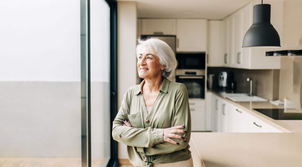Confident older woman with gray hair standing arms crossed in a modern kitchen, smiling thoughtfully near a large window