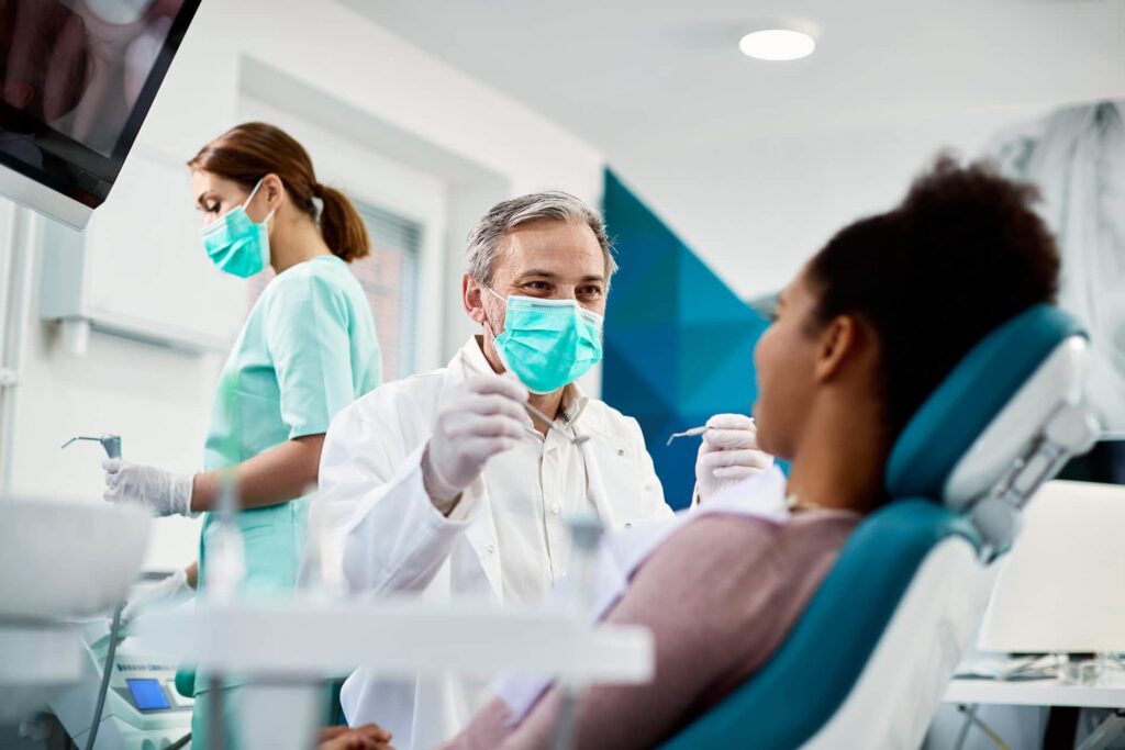 Dentist in white coat and mask examining a female patient in a dental chair while an assistant prepares tools nearby
