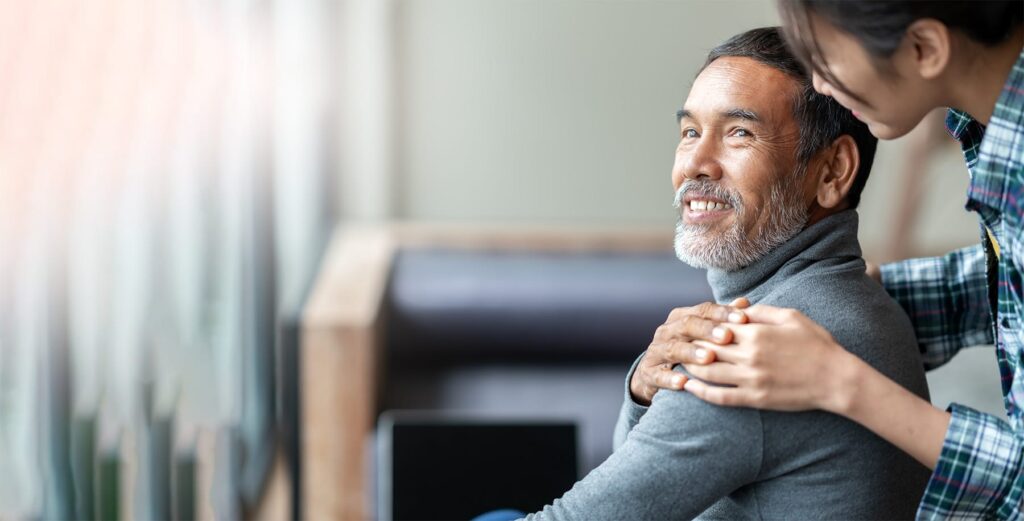 Smiling older man with gray beard receives a comforting hand on his shoulder from a younger woman indoors