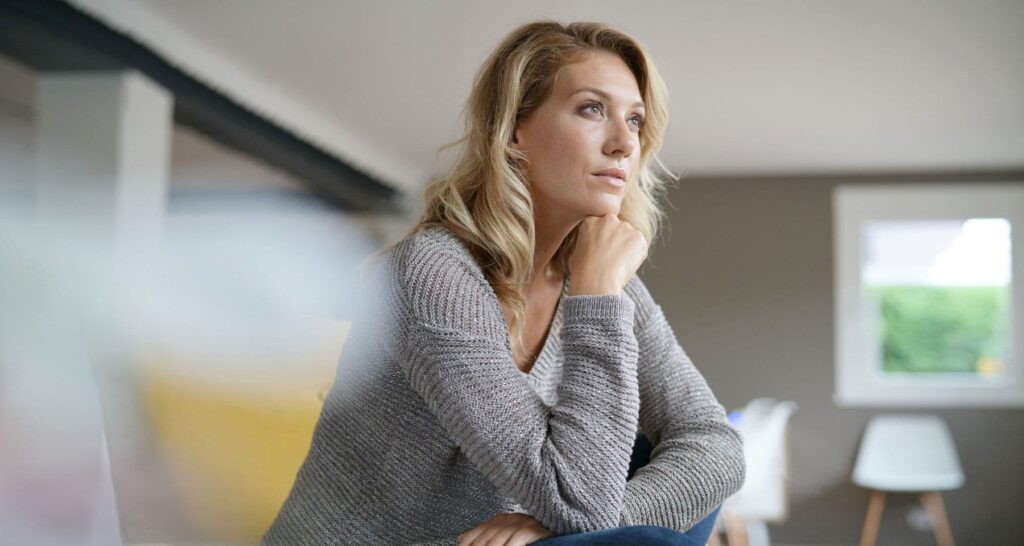 Thoughtful woman in gray sweater sitting at home, resting chin on hand with a contemplative expression