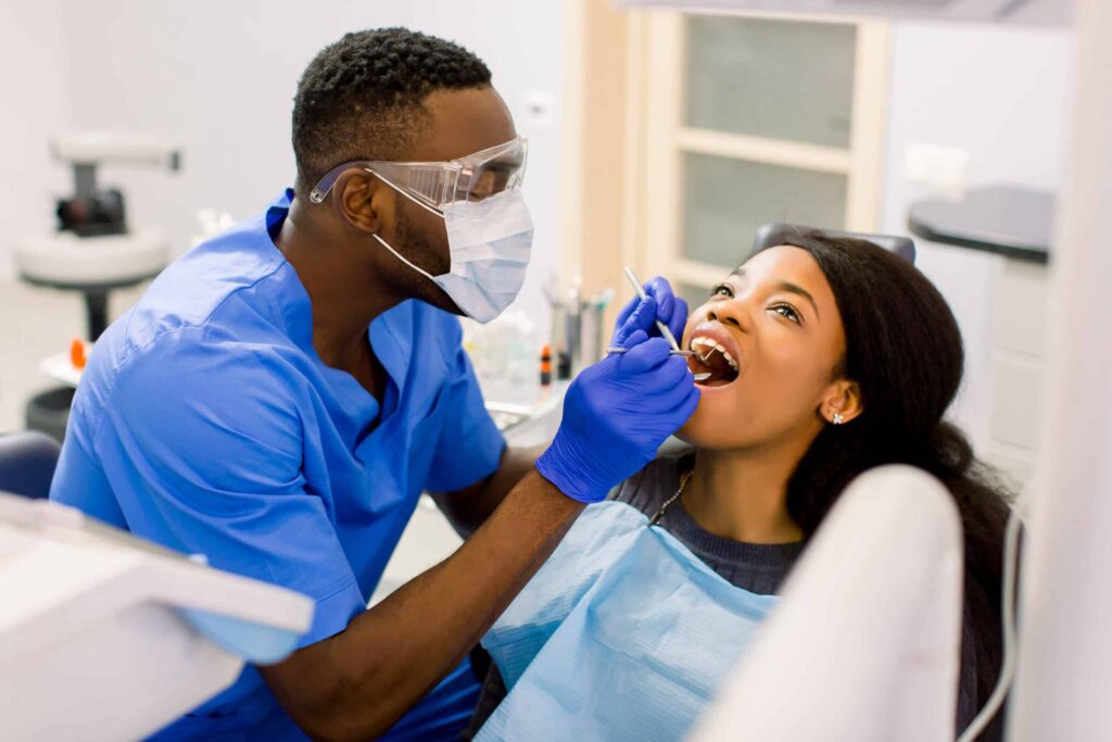 Dentist in blue scrubs and protective gear examining a female patient's teeth during a dental office procedure