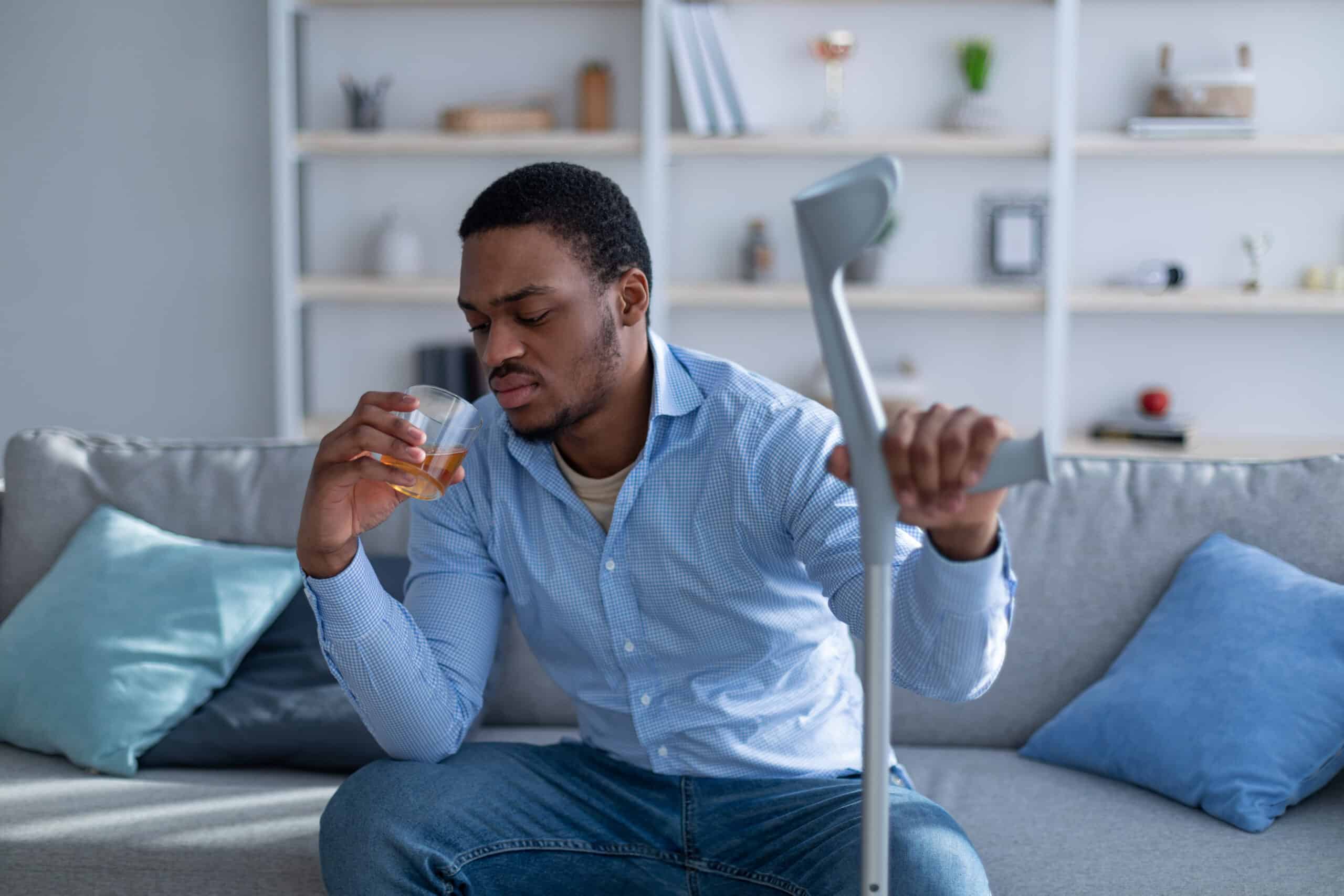 Injured man sitting on couch holding a crutch and a glass of alcohol, showing signs of distress after an accident