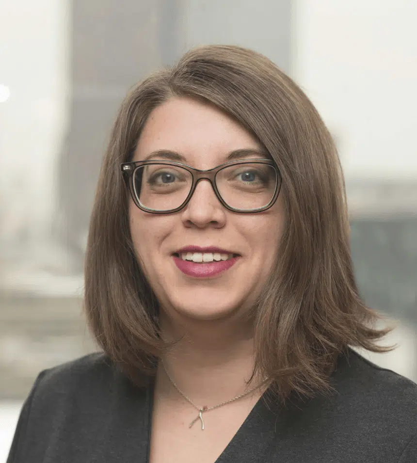 Female attorney in dark blazer and glasses smiling in professional headshot with blurred city office background